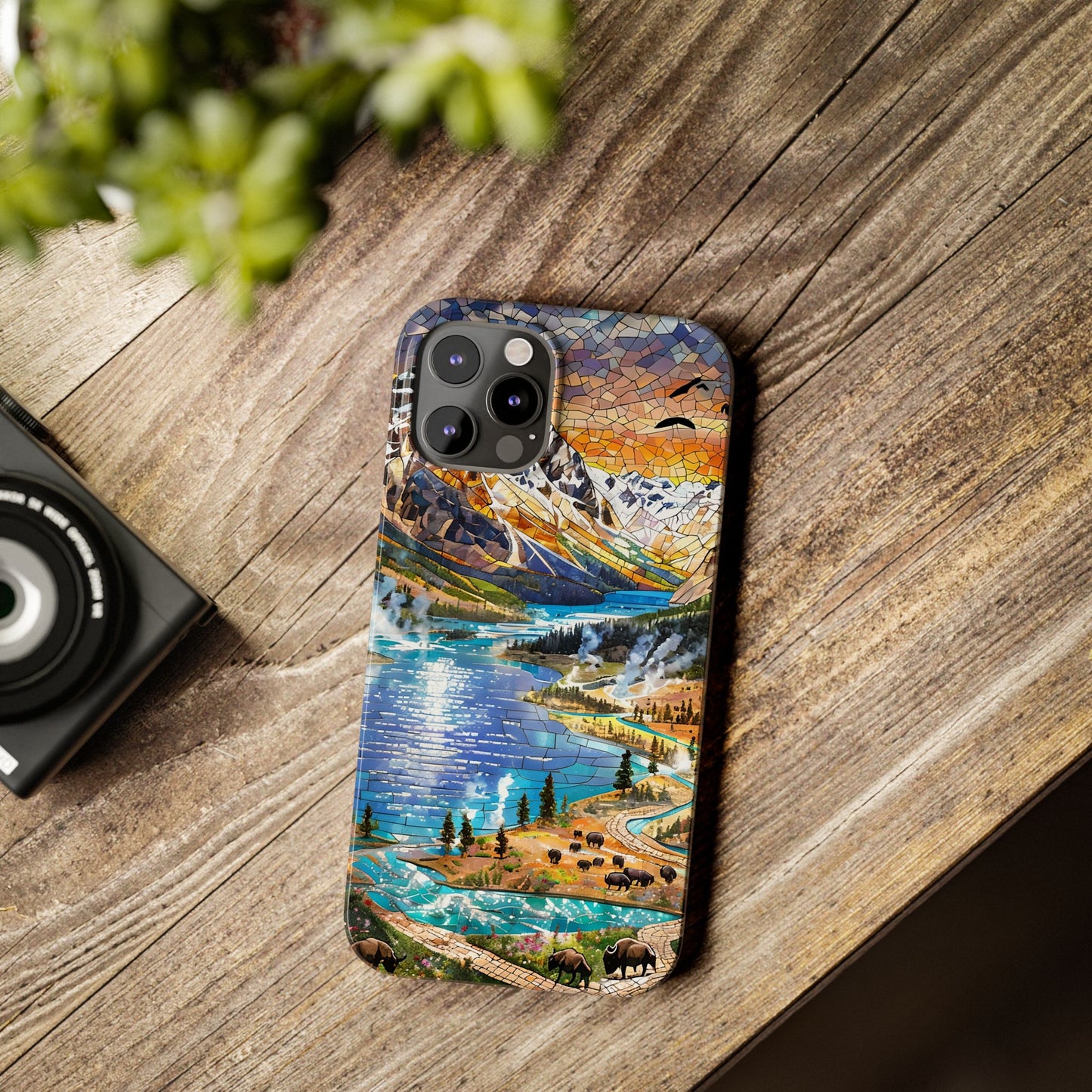 May include: A phone case featuring a mosaic-style landscape design with a lake, mountains, and a sunset. The case has a blue, orange, and green color palette. The phone case has a camera module with three lenses.
