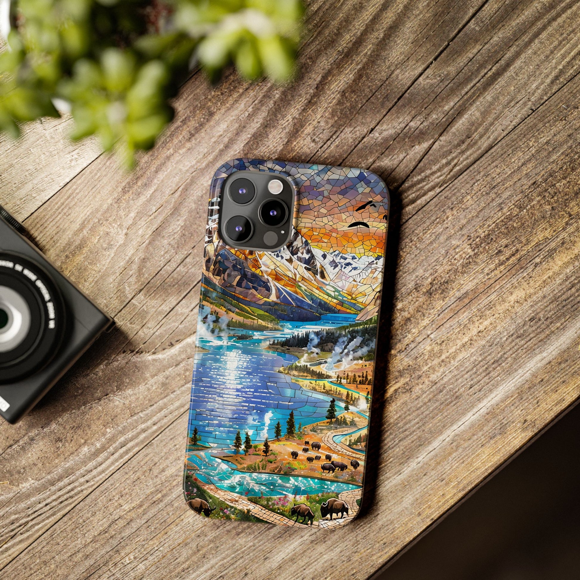 May include: A phone case featuring a mosaic-style landscape design with a lake, mountains, and a sunset. The case has a blue, orange, and green color palette. The phone case has a camera module with three lenses.